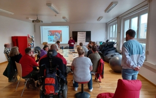 Mehrere Menschen zum Teil im Rollstuhl sitzen um einen Tisch und schauen zur angehenden Bildungsfachkraft Franziska Reschke, die den Veranstaltungsablauf erklärt.